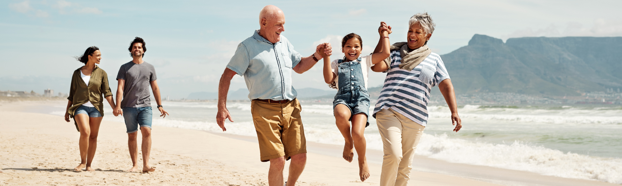 Familie ist gemeinsam am Strand unterwegs. Die Großeltern heben ihr Enkelkind gemeinsam in die Luft und lachen dabei.